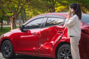 red vehicle partially crushed by accident and young woman on the phone, calling insurance provider Bundle Bee Agency